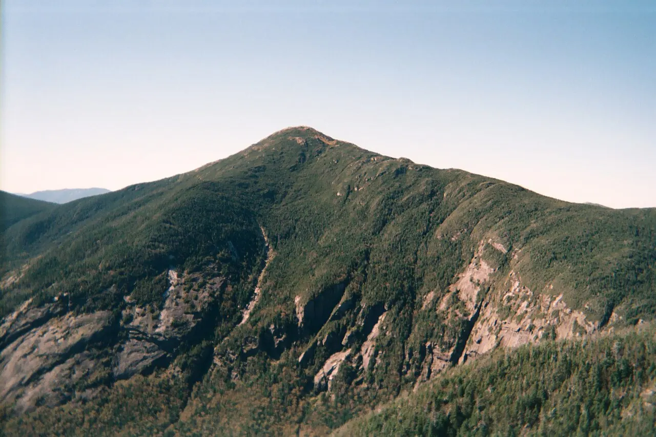 Mount Marcy and the surrounding forested Adirondack High Peaks under a partly cloudy sky, seen from the rocky summit of Mount Haystack.