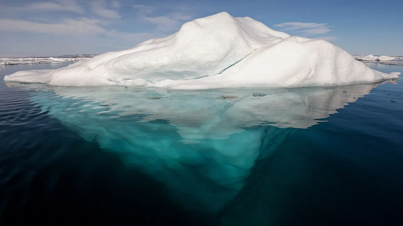 An iceberg floating in calm Arctic water with most of its mass visible below the surface, only a small portion rising above the waterline.