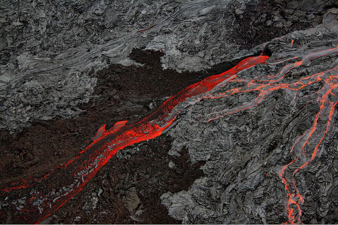 Aerial view of glowing molten lava flowing over older dark hardened lava in Hawaii, showing smooth ropy pahoehoe and rough blocky aa textures side by side.